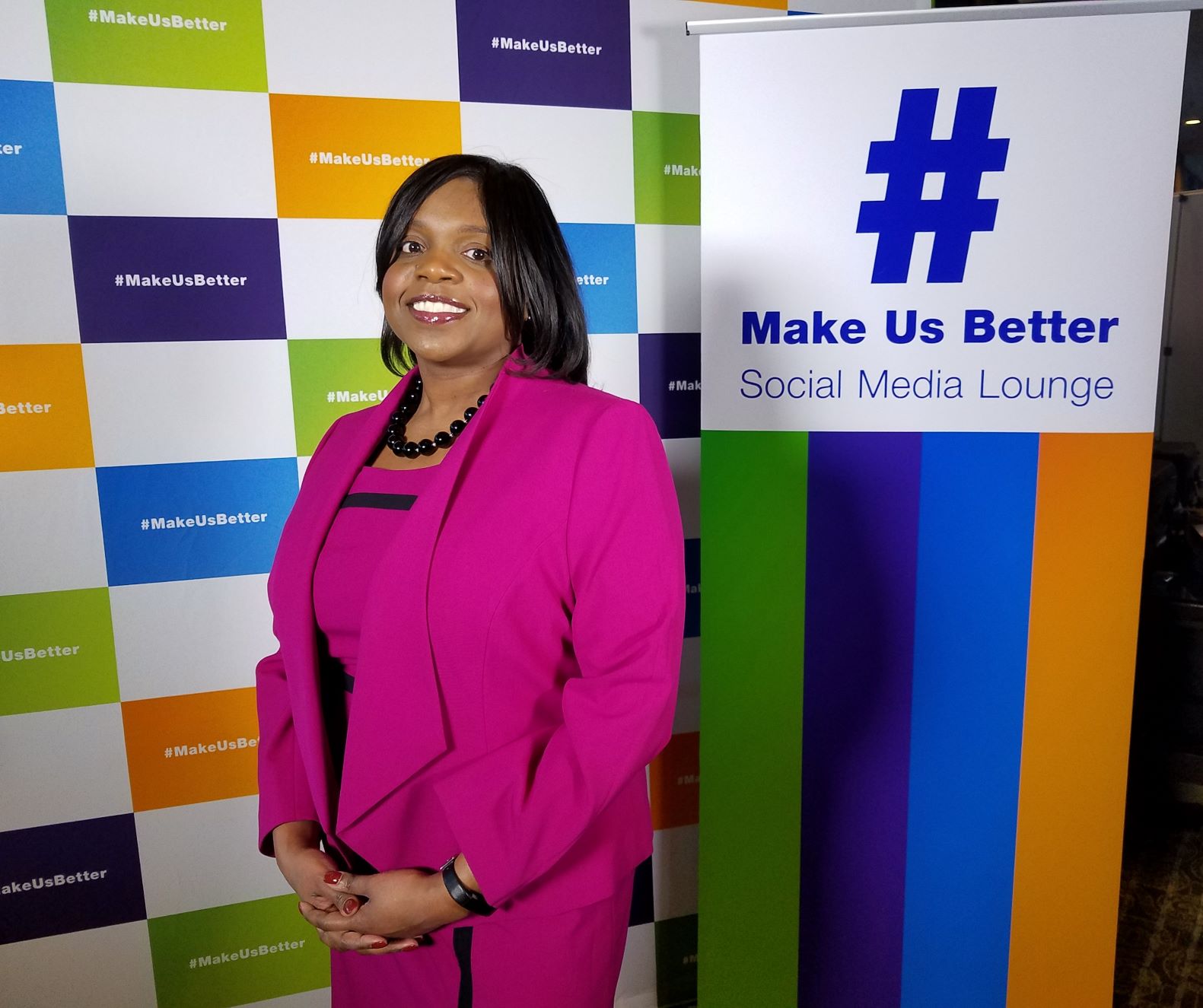 Kimberly poses in front of colorful backdrop and signage, including sign that reads, “# Make Us Better”.